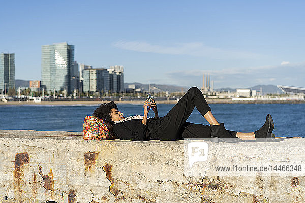 Spanien  Barcelona  schwarz gekleidete Frau  die mit einem Smartphone an einer Wand vor dem Meer liegt