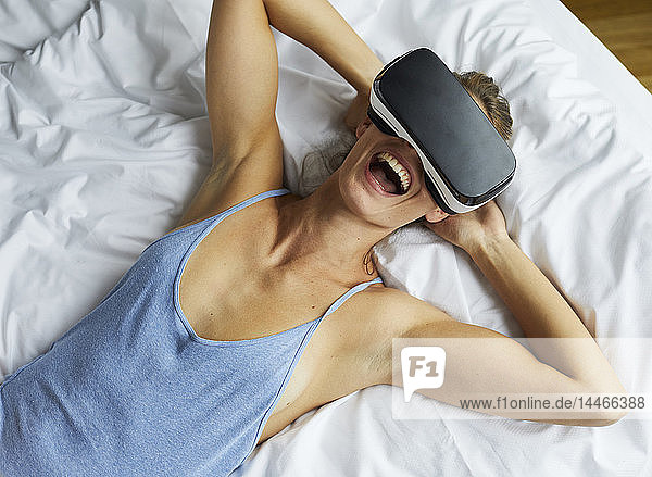 Lachende junge Frau mit VR-Brille im Bett liegend