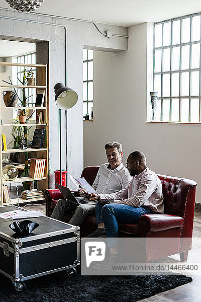 Two businessmen using laptop and discussing documents on sofa in loft office
