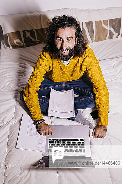 Portrait of happy young man working in bed at home with laptop and documents