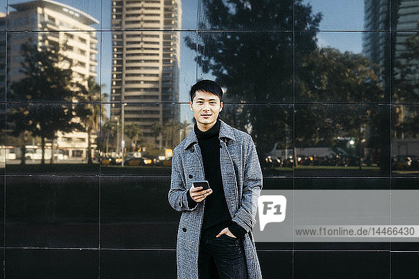 Spanien  Barcelona  Porträt eines jungen Mannes mit Mobiltelefon in schwarzem Rollkragenpullover und grauem Mantel