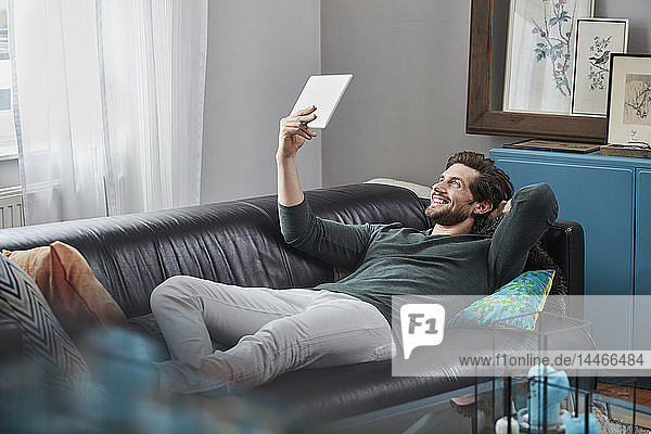 Lächelnder Mann mit Tablette  der zu Hause auf der Couch liegt