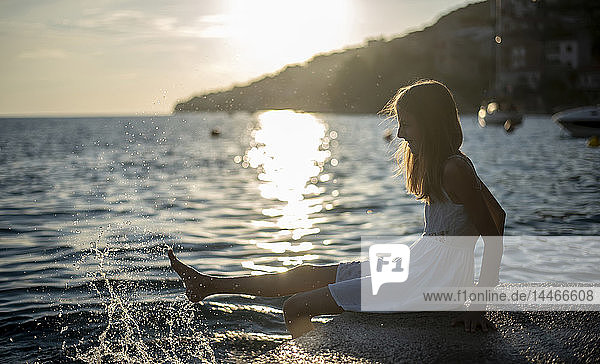 Kroatien  Lokva Rogoznica  Mädchen sitzt bei Sonnenuntergang am Wasser und spritzt mit Wasser
