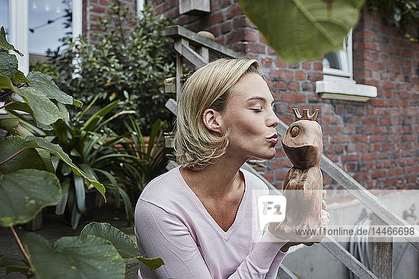 Junge Frau sitzt auf einer Treppe und küsst einen hölzernen Froschprinzen