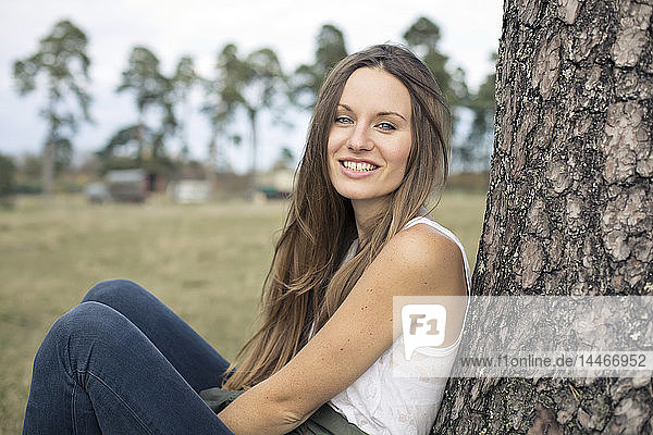 Porträt einer lächelnden jungen Frau  die sich an einen Baumstamm lehnt