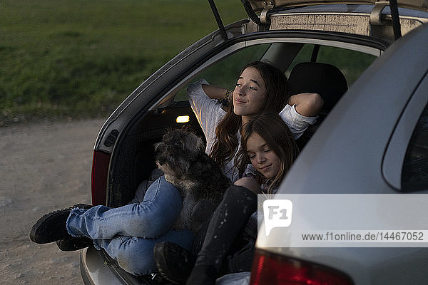 Zwei Schwestern im geöffneten Kofferraum eines geparkten Autos beim Sonnenuntergang
