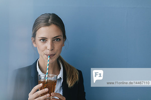 Porträt einer jungen Geschäftsfrau vor blauem Hintergrund  die mit einem Strohhalm Eistee trinkt