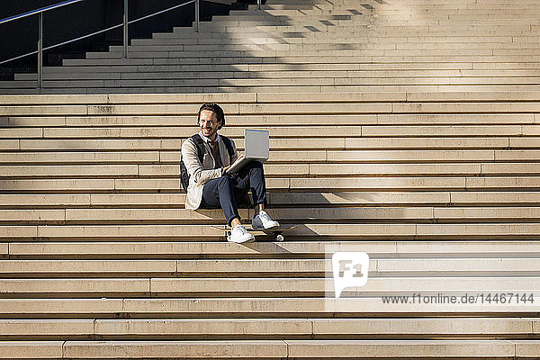 Laughing man with backpack and skateboard sitting on stairs using laptop