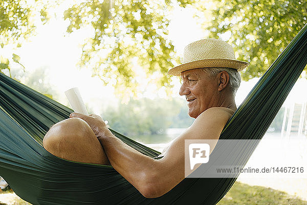 Älterer Mann mit Strohhut entspannt sich in Hängematte am Seeufer und liest Buch