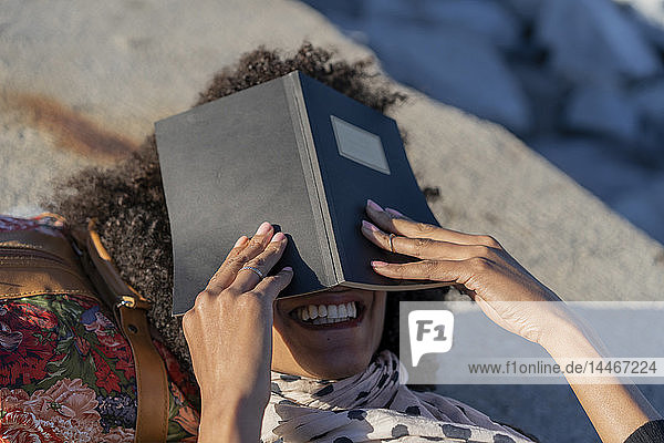 Lachende Frau  die an einer Wand liegt und ihr Gesicht hinter einem Notizbuch verbirgt
