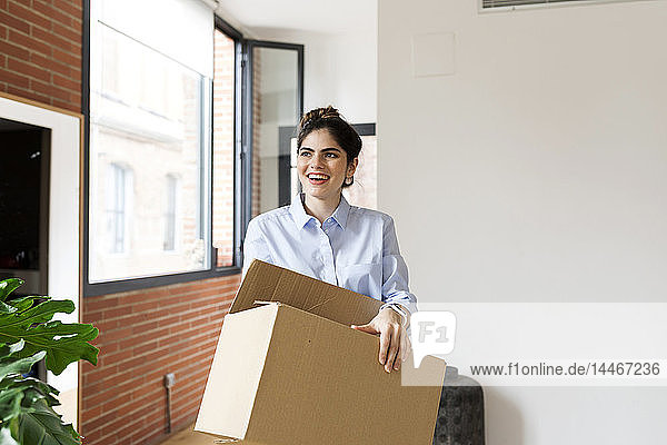 Smiling young woman carrying cardboard box in new apartment