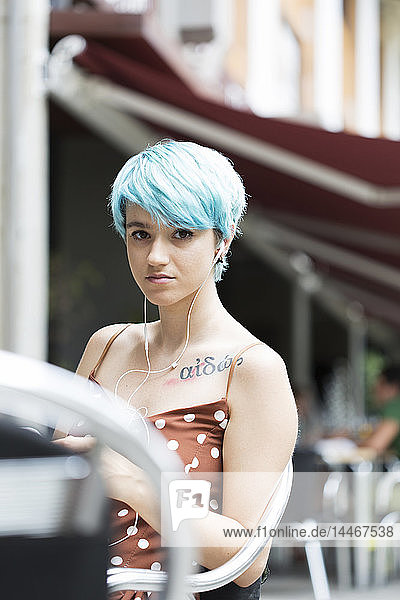 Portrait of young woman with blue dyed hair sitting in a pavement cafe listening music with earphones