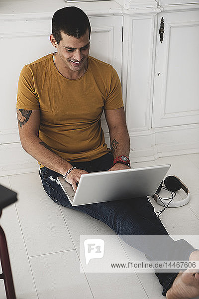 Lächelnder junger Mann sitzt mit Laptop auf dem Boden