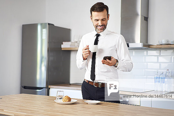 Mann  der vor der Arbeit in der Küche steht  Kaffee trinkt und auf sein Telefon schaut