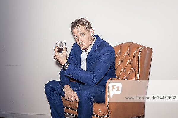 Porträt eines blonden jungen Mannes in blauem Anzug auf einem Lederstuhl sitzend mit einem Getränk