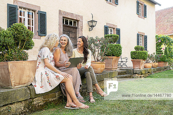 Drei glückliche Frauen unterschiedlichen Alters sitzen mit Tablette im Garten