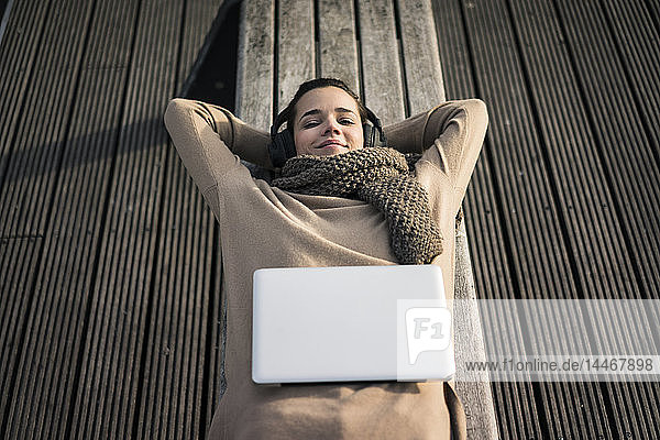 Porträt einer lächelnden Frau  die auf einer Bank liegt und mit schnurlosen Kopfhörern Musik hört