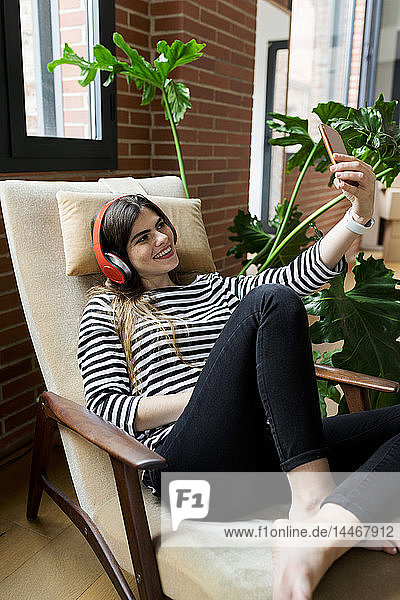 Junge Frau  die zu Hause im Sessel sitzt und mit Kopfhörern Musik hört und sich ein Selfie