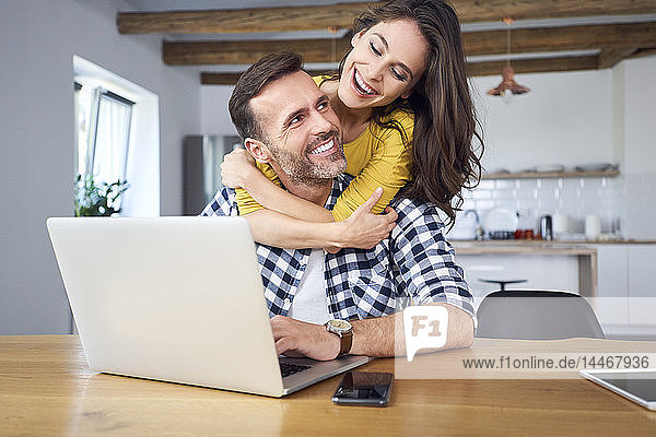 Glückliches Paar sitzt am Esstisch  umarmt sich  benutzt einen Laptop