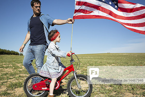 Mann und Tochter mit Fahrrad und amerikanischer Flagge auf Feld in entlegener Landschaft