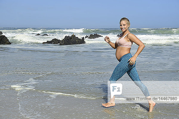 Lächelnde schwangere Frau joggt am Strand