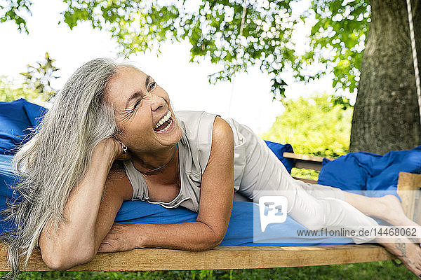Lachende Frau mit langen grauen Haaren liegt auf einem Bett im Garten