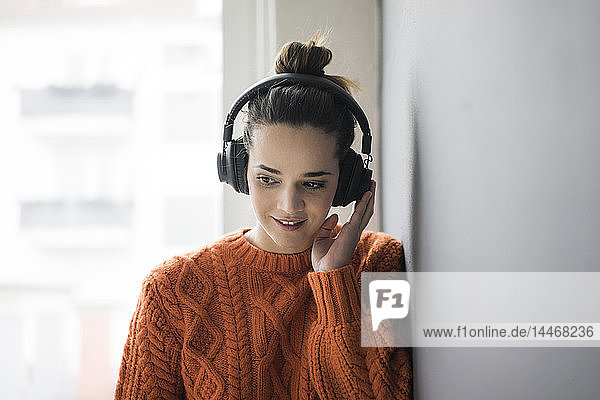 Porträt einer lächelnden Frau in orangem Strickpullover  die mit schnurlosen Kopfhörern Musik hört