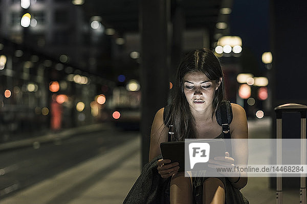 Portrait of young woman with headphones and tablet waiting at station by night