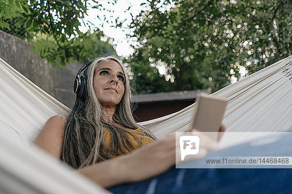 Porträt einer Frau mit Mobiltelefon  die in der Hängematte im Garten liegt und mit Kopfhörern Musik hört