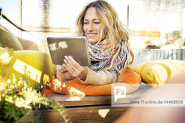 Porträt einer lächelnden  blonden  reifen Frau  die sich im Wintergarten mit einem digitalen Tablet entspannt