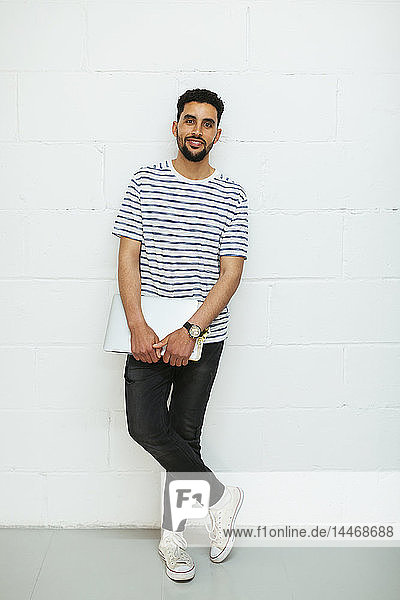 Portrait of smiling young man with laptop standing at brick wall