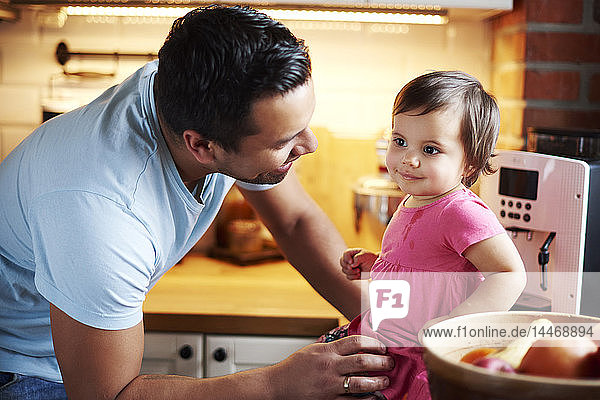 Smiling father looking at baby girl sitting on counter in kitchen at home