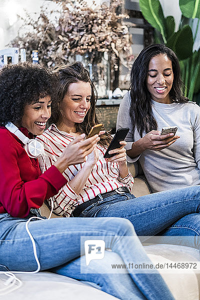 Three smiling women sitting on couch using cell phones