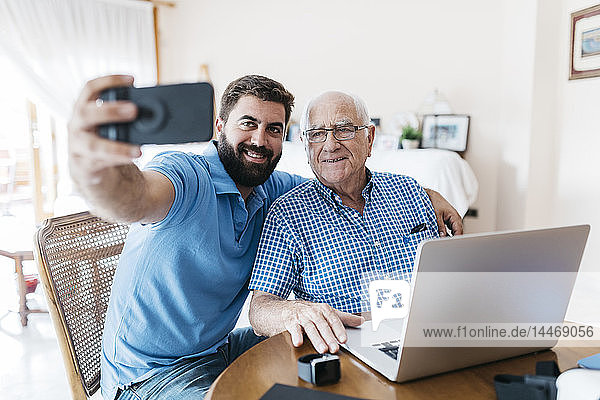 Porträt eines erwachsenen Enkels und seines Großvaters  die sich zu Hause mit einem Smartphone selbstständig machen