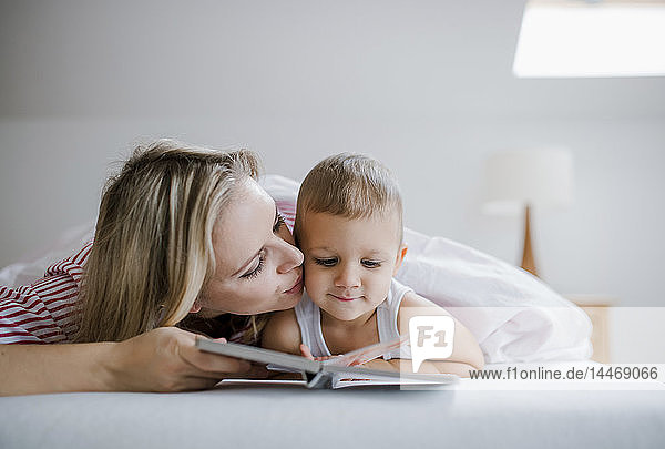 Mother kissing toddler son lying in bed at home reading a book