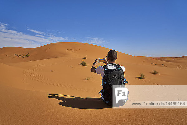 Morocco  man with backpack sitting on desert dune taking picture with smartphone