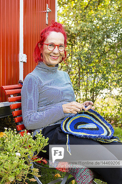 Porträt einer lächelnden älteren Frau mit rot gefärbten Haaren  die im Garten vor einem roten Wohnwagen sitzt und häkelt