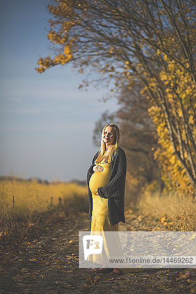 Lächelnde schwangere Frau steht im Herbst auf einem Waldweg neben einem Spargelfeld