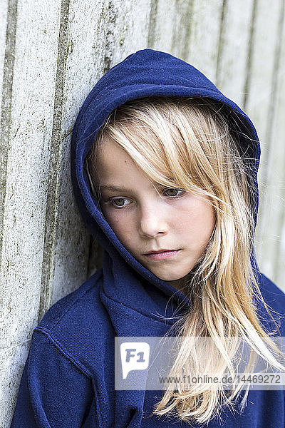 Bildnis eines traurigen blonden Mädchens mit blauer Kapuzenjacke  das an einer Holzwand lehnt