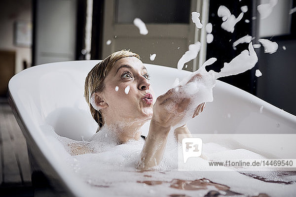 Blond woman taking bubble bath blowing foam in the air