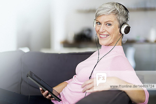 Porträt einer glücklichen  reifen Frau  die mit einem digitalen Tablet auf der Couch sitzt und mit Kopfhörern Musik hört