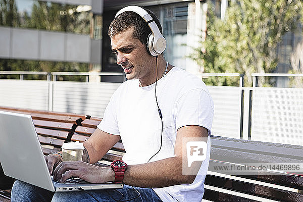 Young man sitting on a bench wearing headphones and using laptop