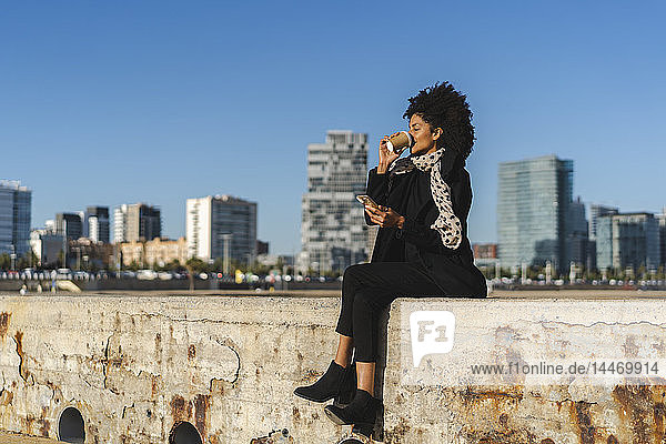 Spanien  Barcelona  Frau mit Smartphone sitzt an einer Wand und trinkt Kaffee zum Mitnehmen