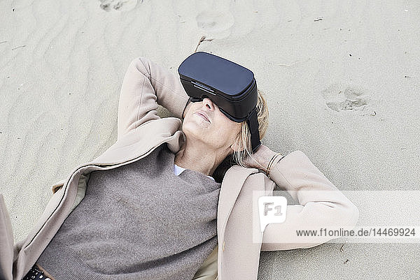 Spain  Menorca  senior woman lying on the beach in winter using VR glasses