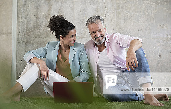 Casual businessman and businesswoman sitting on artificial turf in a loft sharing laptop