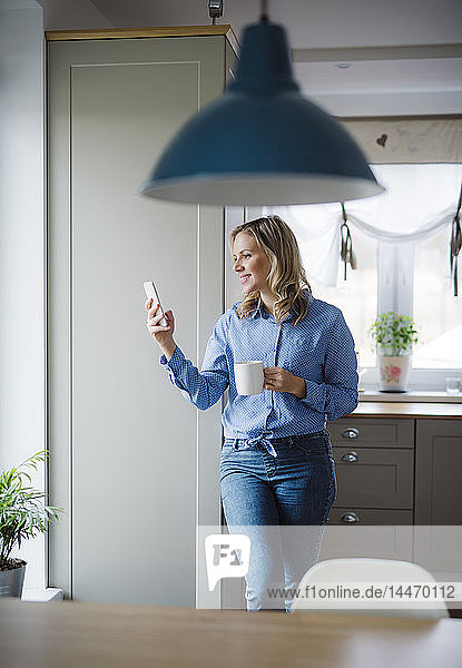 Lächelnde Frau hält zu Hause eine Tasse Kaffee und ein Smartphone