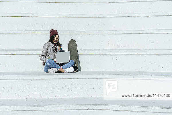Junge Frau mit Skatoboard sitzt mit Laptop auf einer Treppe im Freien