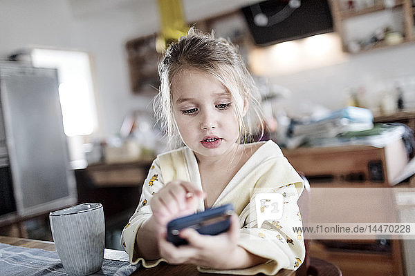 Portrait of little girl sitting at breakfast table in the kitchen using smartphone
