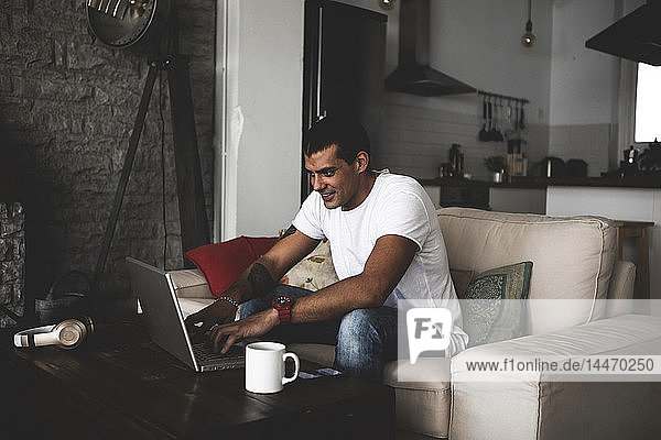 Smiling young man sitting on sofa at home using laptop