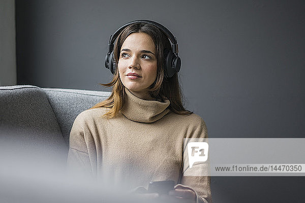 Porträt einer Frau mit Smartphone und schnurlosen Kopfhörern  die sich auf der Couch entspannt und aus dem Fenster schaut